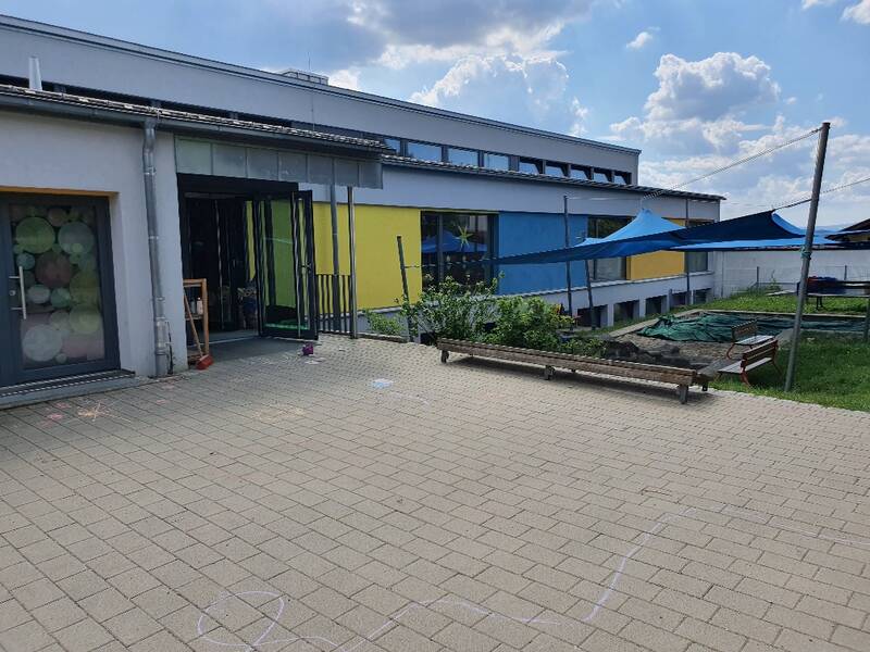 Flachdachgeäude, bunte Außenwände wechseln sich mit großen Glasfronten ab, rechts im Bild ein Spielbereich mit Sandkasten, der mit einem bunten Sonnensegel überspannt ist. der Vorplatz ist mit bunter Malkreide bemalt.