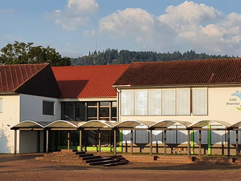 Das Foto zeigt ein modernes Schulgebäude mit rotem Ziegeldach und einem Logo, das auf der Fassade angebracht ist, umgeben von Bäumen und einem blauen Himmel im Hintergrund. Im Vordergrund der Schulhofplatz mit einem überdachten Weg zum Eingang.  In der Mitte des Weges steht eine Sitzbank. 