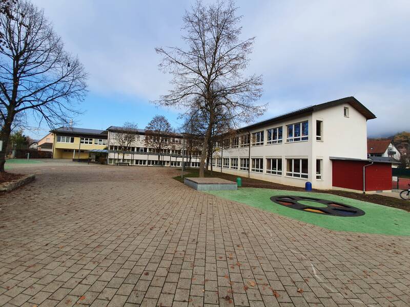 Ein Schulgebäude mit mehreren Flügeln, weißen Wänden und einem flachen Dach ist umgeben von einem Pausenhof mit Spielmarkierungen und Bäumen bei herbstlichem Tageslicht.