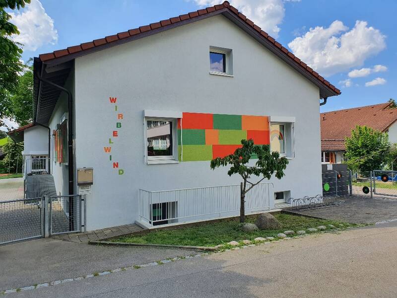 Ein weißes zweistöckiges Haus mit farbigen Quadraten an der Fassade und der Aufschrift "WIRBELWIND" an einer Seite unter blauem Himmel.