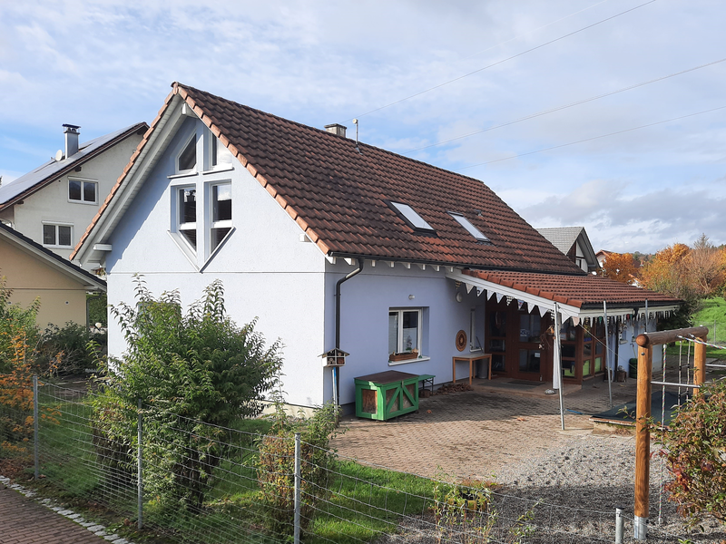 Ein zweistöckiges Haus mit rotem Ziegeldach, umgeben von einem Garten und einem Zaun, steht bei sonnigem Wetter neben anderen Häusern.