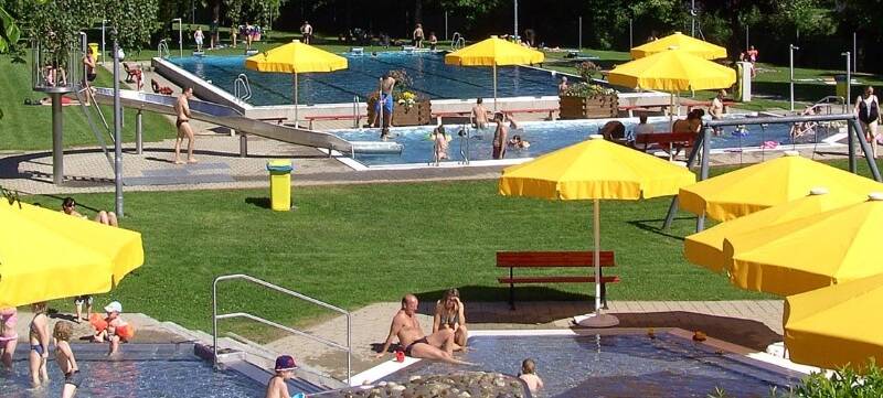 Freibad von Bäumen und Hecken umgeben, mit großzügigen Liegeflächen, gelbe Sonnenschirme verteilt um mehrere Schwimmbecken und Sitzgelegenheiten, eine Rutschbahn führt in ein Schwimmbecken, an mehreren Plätzen und im Wasser tummeln sich Badebesucher bei blauem Himmel und Sonnenschein. 