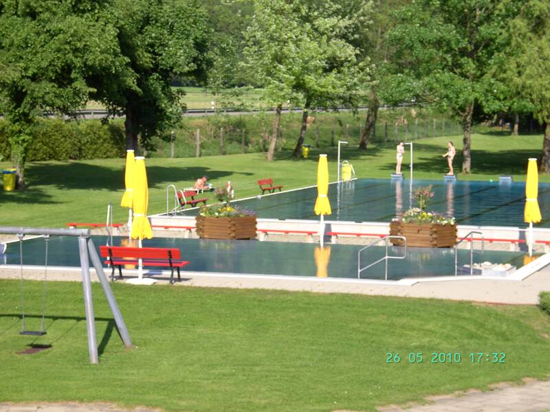 Freibad von Bäumen und Hecken umgeben, mit großzügigen Liegeflächen, gelbe geschlossene Sonnenschirme verteilt um das Schwimmerbecken und Sitzgelegenheiten, eine Schaukel im Vordergrund, zwei Kinder auf den Startblöcken