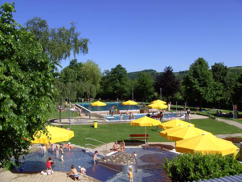 Freibad von Bäumen und Hecken umgeben, mit großzügigen Liegeflächen, gelbe Sonnenschirme verteilt um mehrere Schwimmbecken und Sitzgelegenheiten, eine Rutschbahn führt in ein Schwimmbecken, an mehreren Plätzen und im Wasser tummeln sich Badebesucher bei blauem Himmel und Sonnenschein. 