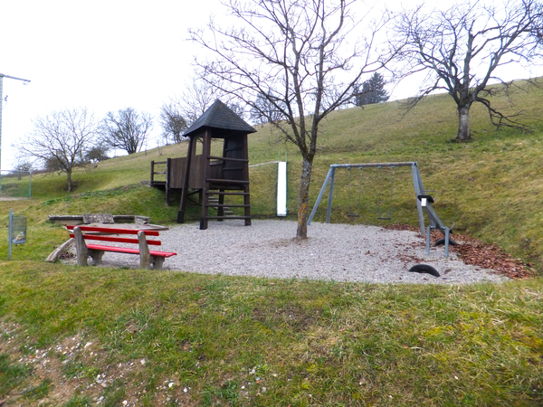 Spielplatz im Herbst mit einem Sandkasten, einem Kletterturm, einer Schaukel und einer Wippe, im Vordergrund steht eine rote Sitzbank