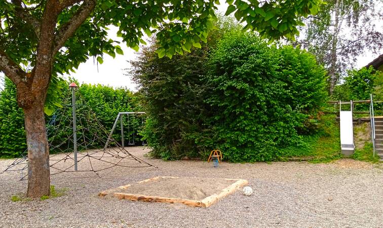 Ein Spielplatz mit Kletterpyramide, Rutsche, Sandkasten, Schaukel und Wipp-Tier, umrandet mit hohen grünen Hecken, links steht ein großer Baum