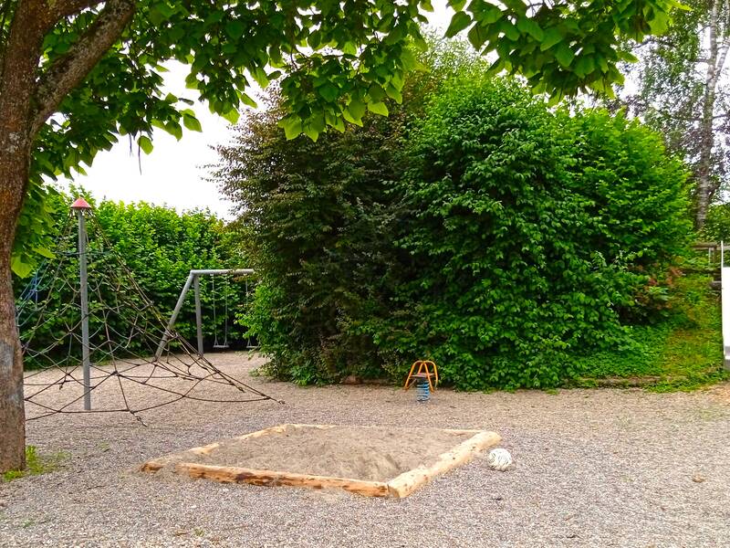 Ein Spielplatz mit Kletterpyramide, Rutsche, Sandkasten, Schaukel und Wipp-Tier, umrandet mit hohen grünen Hecken, links steht ein großer Baum