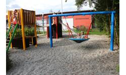 Spielplatz, zwei Kinder auf einer großen runden Schaukel,  daneben ein Kletterturm mit einer Rutsche an der anderen Seite. Auch hier spielende Kinder. Im Hintergrund die Vorderseite des Dorfgemeinschaftshauses