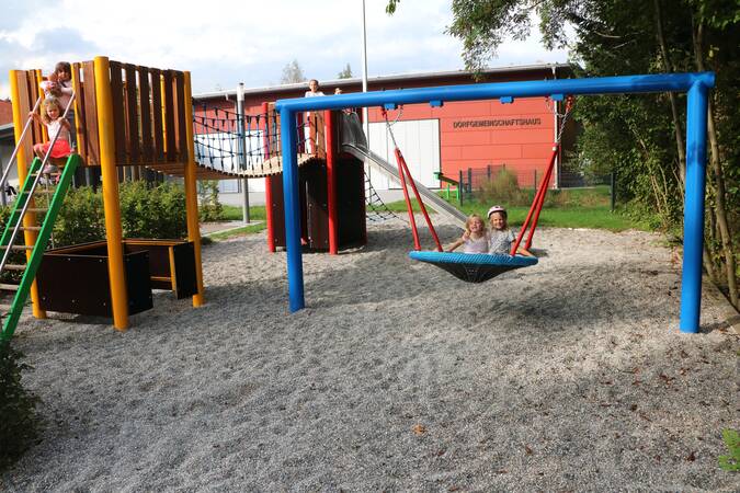 Spielplatz, zwei Kinder auf einer großen runden Schaukel,  daneben ein Kletterturm mit einer Rutsche an der anderen Seite. Auch hier spielende Kinder. Im Hintergrund die Vorderseite des Dorfgemeinschaftshauses