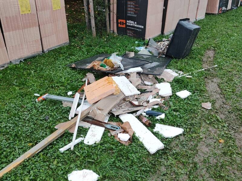 Auf einer Wiese liegen Bauschutt und Styroporstücke vor gestapelten Baumaterialien neben einem Baum.