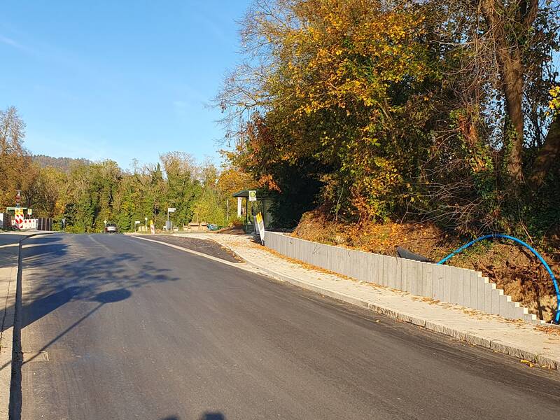 Eine neu asphaltierte Straße führt an herbstlich gefärbtem Gehölz entlang, mit einem Baustellenschild und einer Absperrung auf dem Gehweg.