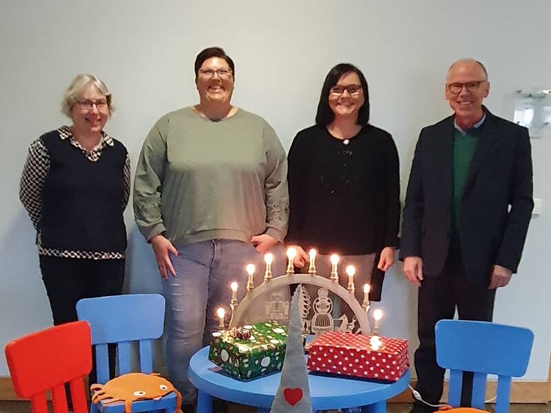 4 Personen stehen nebeneinander, davor ein kleiner Kindertisch mit bunten Kinderstühlen darum, ein Weihnachtsmann aus Stoff seht vor dem Tisch, auf dem Tisch ein Lichterbogen aus Holz und bunte Geschenke