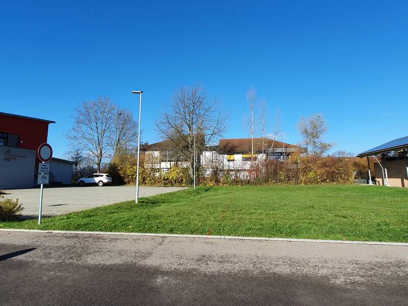 große Wiese, linker Seite das rote Feuerwehrgebäude, hin Hintergrund hinter einer grünen Hecke das Schulgebäude, rechts das Bauhofgebäude, strahlend blauer Himmel