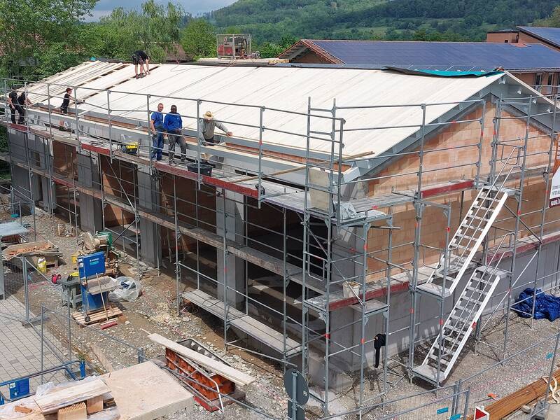 ein zweistöckiges eingerüstetes Gebäude im Rohbau, viele Arbeiter um und auf dem Gebäude, Dachdecker auf dem Dach, 