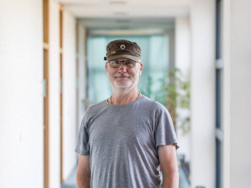 Mann mit brauner Mütze, Brille, Kinnbart und grauem Shirt steht in einem langen Hellen Flur