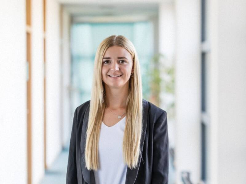 Frau mit langen Blonden Haaren, schwarzem Blazer, weißem Shirt steht in einem langen Hellen Flur