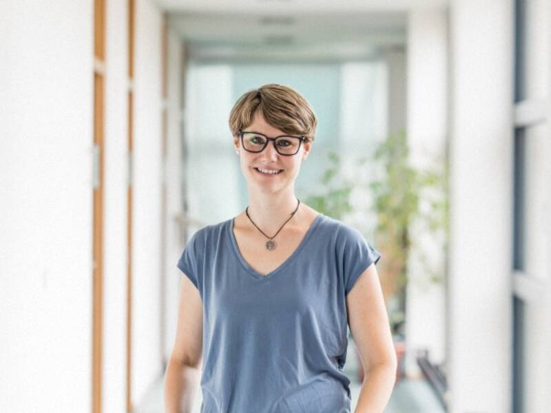 Frau mit kurzen braunen Haaren, Brille, Kette, blauen Kurzarmshirt steht in einem langen Hellen Flur