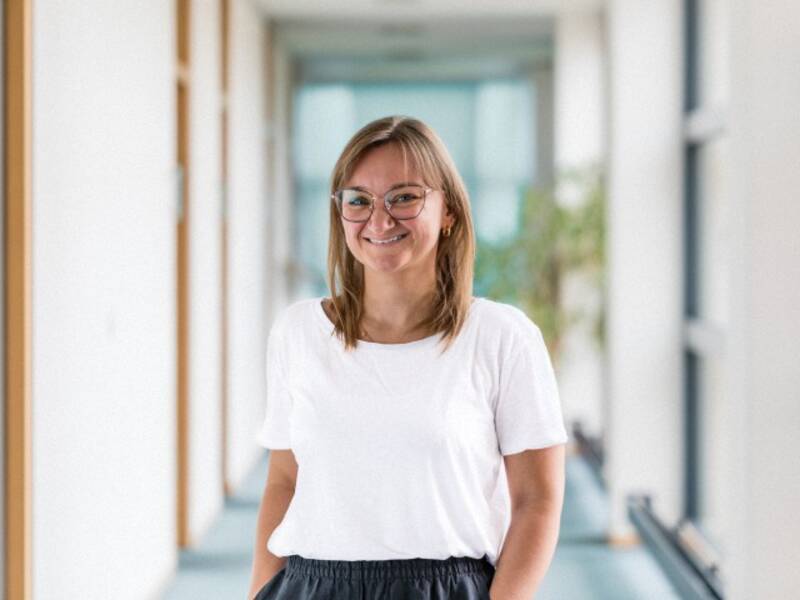 Frau mit schulterlagen braunen Haaren, Brille, weißem Shirt steht in einem langen Hellen Flur