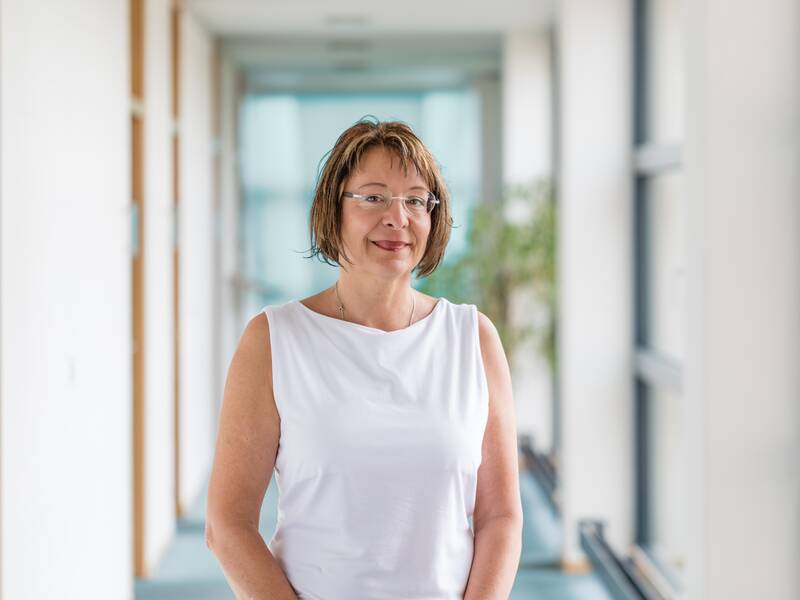 Frau mit kurzem braunem Haar, Brille und weißen Shirt  steht in einem langen Hellen Flur