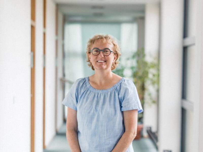 Eine Frau mit kurzen blonden Locken, Brille und hellblauem Kurzarmshirt steht in einem langen Hellen Flur