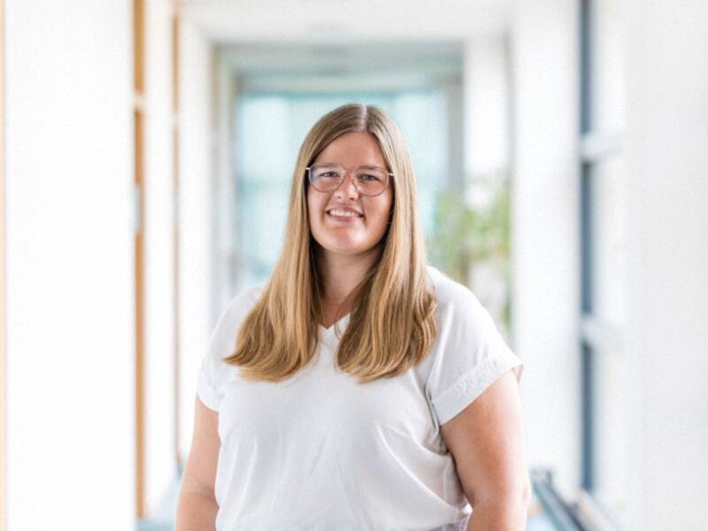 Eine Frau mit dunkelblonden Haaren, einer Brille und einem weißen Oberteil vor verschwommenen Hintergrund. 