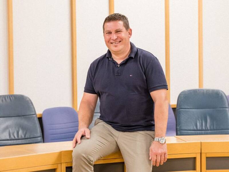 Mann mit dunklen kurzen Haaren, blauem Poloshirt und heller Hose lehnt an einem halbrunden Holztisch mit dunklen Lederstühlen im Hintergrund