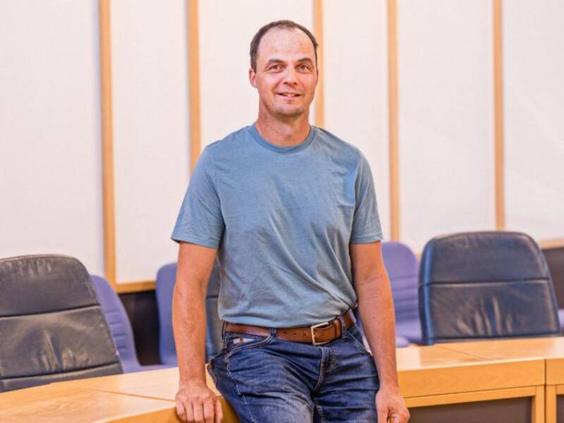 Mann im blauen T-Shirt, Jeans, Gürtel und dunklen Haaren mit Stirnglatze steht vor einem halbrunden Holztisch mit dunklen Stühlen im Hintergrund