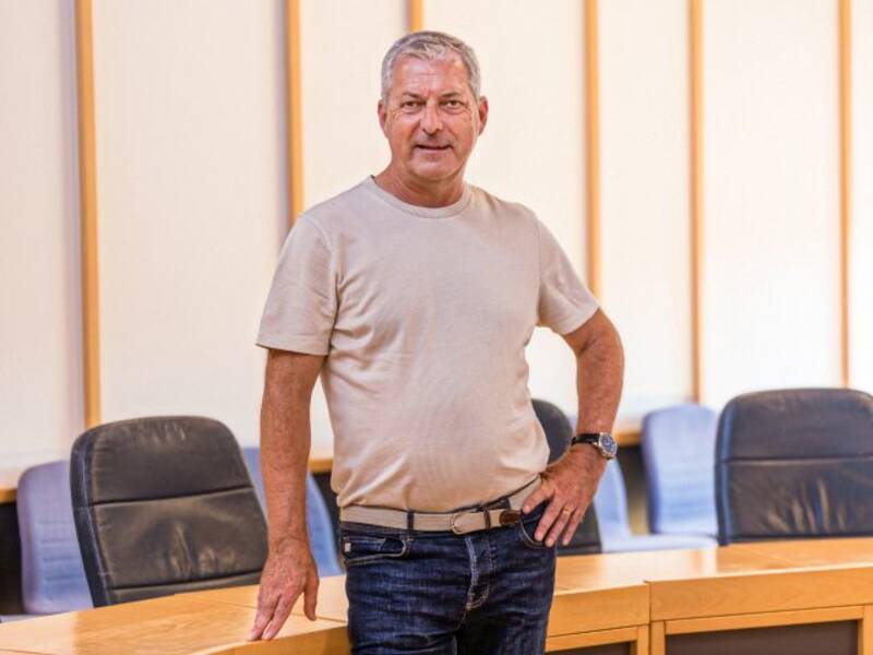 Mann mit beigem T-Shirt, Jeans, Gürtel und hellen, kurzen Haaren steht vor einem halbrunden Holztisch mit dunklen Lederstühlen im Hintergrund