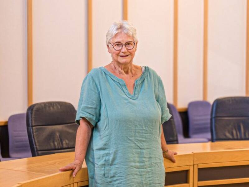 Frau mit hellen Haaren und blaugrüner Bluse und Brille steht vor einem halbrunden Holztisch mit dunklen Lederstühlen im Hintergrund