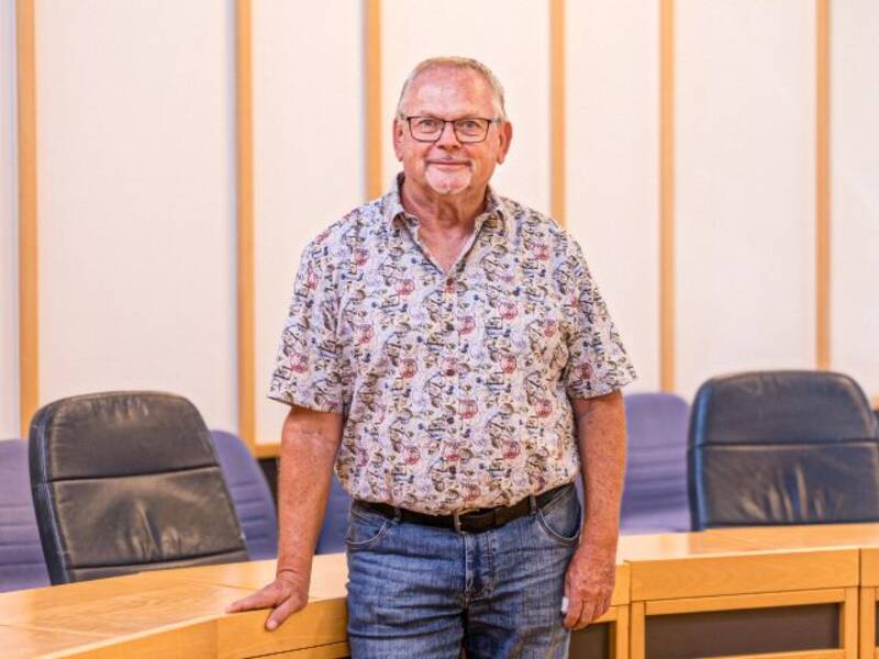 Mann mit grauen Haaren, Brille, Kinnbart, bunt gemustertem Kurzarmhemd, Jeans und Gürtel steht vor einem halbrunden Holztisch mit dunklen Lederstühlen im Hintergrund