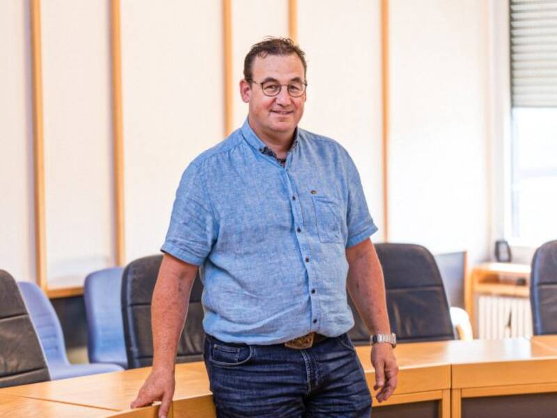 Mann mit blauem Kurzarmhemd, Jeans, Brille und kurzen dunklen Haaren steht vor einem halbrunden Holztisch mit dunklen Lederstühlen im Hintergrund