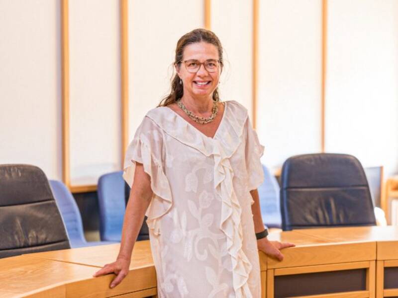 Frau in hellem Kleid, dunklen, langen, zusammengebundenen Haaren, Brille und Kette steht vor einem halbrunden Holztisch mit dunklen Lederstühlen im Hintergrund