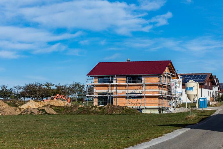 Neu gebautes Haus, das noch eingerüstet ist. Das Haus steht am Ende eines Wohngebiets auf einer grünen Wiese. Der Himmel ist blau.