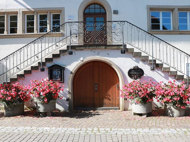 Eingangstür zum Heimatmuseum, darüber ein pyramidenförmiger Treppenaufgang, auf beiden Seiten rosa blühende Blumen